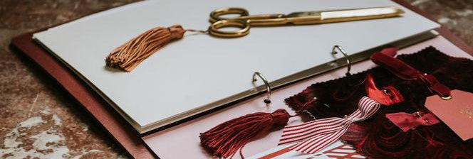 A pair of brass-finished scissors with a tassel on top, placed on an open book or document, surrounded by decorative ribbons and a card.